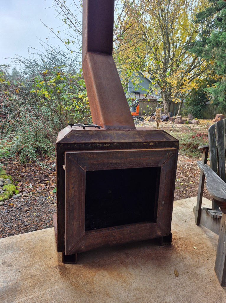 A rusted metal wood stove positioned on a concrete surface surrounded by greenery and trees in the background.